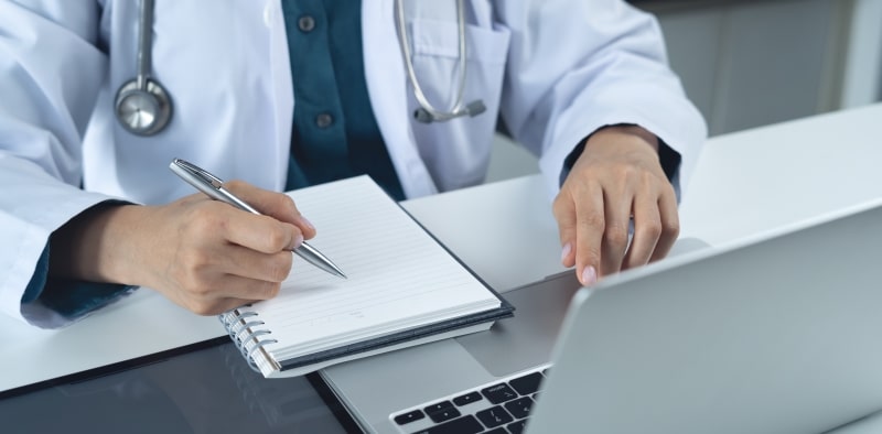 Um médico fazendo anotações em um caderno e computador. Imagem para ilustrar a matéria sobre medicina preventiva para longevidade.