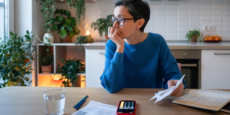 Um mulher reflexiva, segurando contas enquanto está sentada em uma mesa. Ao seu lado está um caderno e em sua frente tem uma calculadora. Imagem para ilustrar a matéria sobre a participação feminina nas finanças.