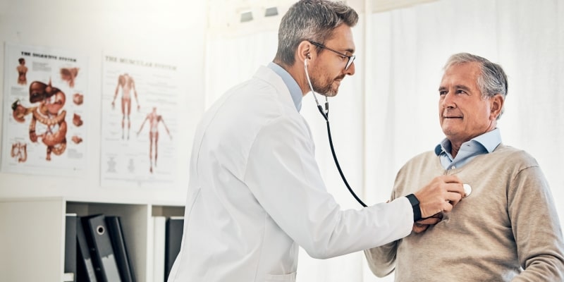 Um homem idoso em uma consulta médico com um médico mais jovem que escuta o seu coração com um estetoscópio. Imagem para ilustrar a matéria sobre a saúde do homem.