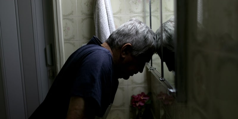 Um homem idoso triste no banheiro, encostando a cabeça no espelho e demonstrando desespero. Imagem para ilustrar a matéria sobre suicídio entre idosos.