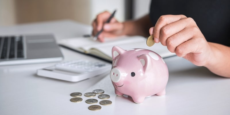 Uma mão feminina colocando moedas em um cofrinho de porco enquanto faz anotações no computador. Imagem para ilustrar a matéria sobre aposentadoria de 5 mil reais.