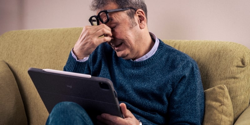 Um homem com cerca de 50 anos com cansaço mental ao usar muito o tablet. Ele está coçando os olhos com o tablet nas mãos enquanto está sentado no sofá.