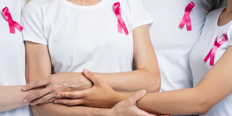 Um grupo de mulheres com blusa branco e um laço de Outubro Rosa se abrançando para ilustrar os desafios na luta contra o câncer de mama.
