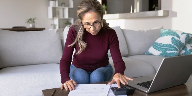 Uma mulher com cerca de 50 anos usando calculadora e computador para fazer um planejamento financeiro. Imagem para ilustrar a matéria sobre hábitos que desperdiçam dinheiro na aposentadoria.