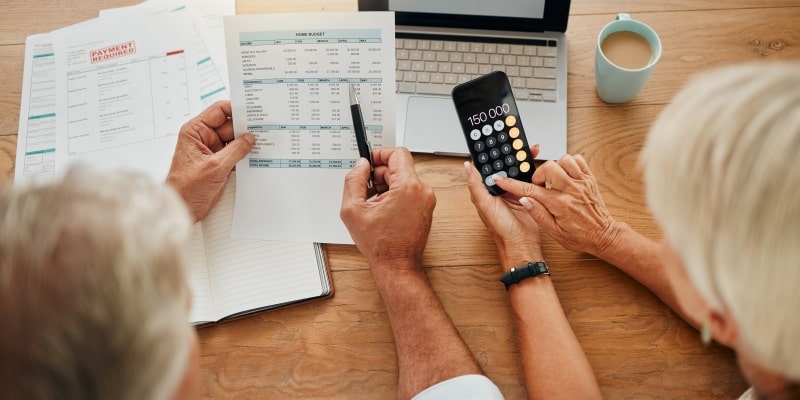 Um casal de idoso fazendo contas em uma calculadora e em um computador, analisando pagamentos. Imagem vista de cima e usada para representar a matéria sobre investir com segurança aos 60 anos.