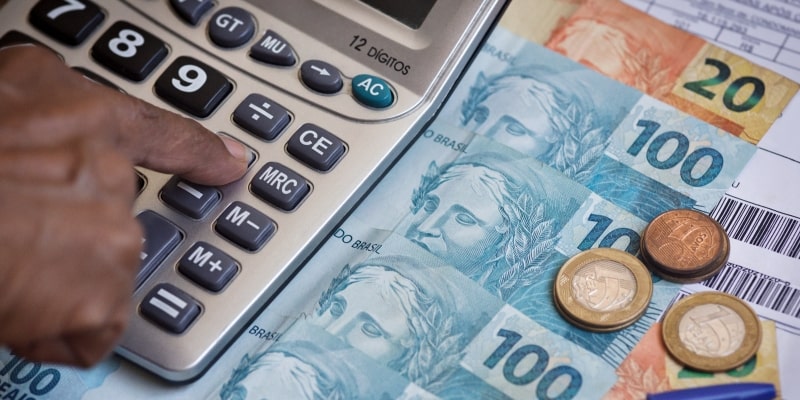 Uma mão negra clicando em uma calculadora que está sobre notas de reais e ao lado de moedas e contas a pagar. Imagem para ilustrar a matéria sobre investir para aposentadoria.