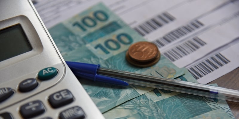 Uma calculadora, notas de 100 reais, moedas, uma caneta e contas a pagar. Imagem para ilustrar a matéria sobre o IPTU 2026.