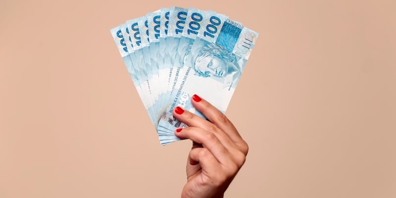 Uma mão feminina segurando notas de 100 reais. Imagem para ilustrar a matéria sobre juros no crédito pessoal.