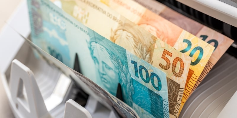 Um contador de dinheiro em notas de reais, ilustrando a matéria sobre o lote extra do PIS-Pasep.