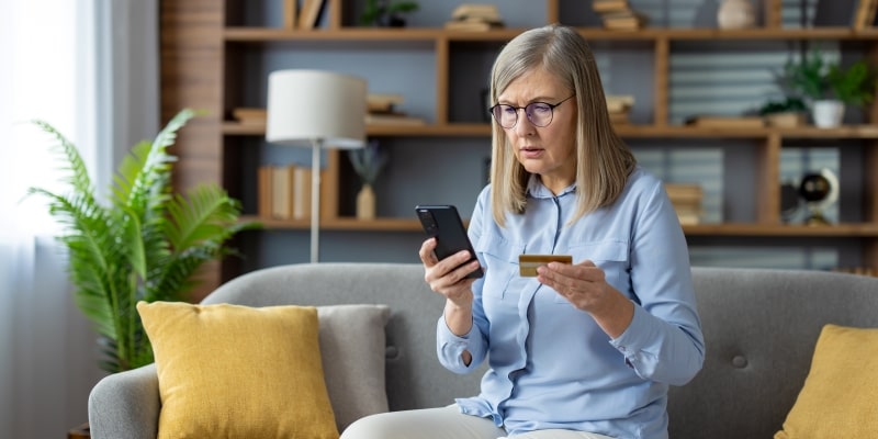 Um mulher segurando um cartão de crédito e olhando com surpresa para o celular, após cair em um golpe de phishing.