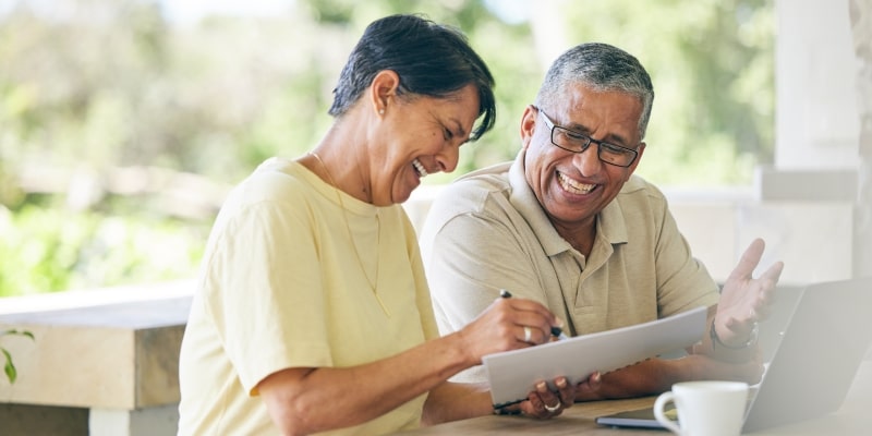 Um homem e uma mulher sorrindo enquanto seguram papéis e olham um computador, fazendo um planejamento financeiro em casal.