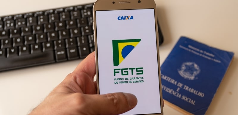 Uma mão segurando um celular com o aplicativo do FGTS. Ao fundo, um teclado de computador e uma carteira de trabalho. Imagem para ilustrar a matéria sobre a recuperação do FGTS.