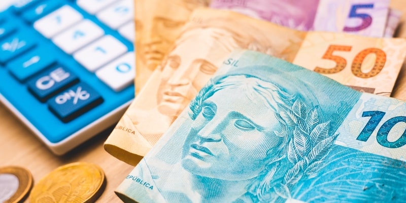Notas de dinheiro, moedas e uma calculadora sobre uma mesa. Imagem para ilustrar a matéria sobre o salário mínimo 2026.