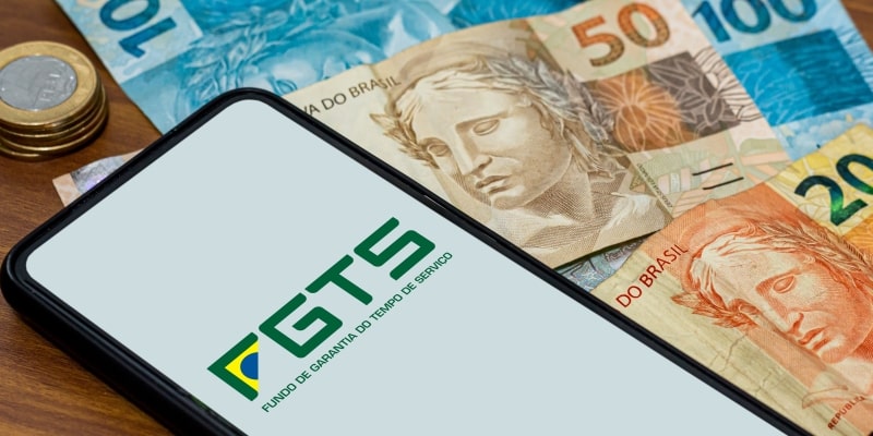 Um celular com o logo do FGTS sobre notas de dinheiro, ilustrando a matéria sobre o saque-aniversário do FGTS.