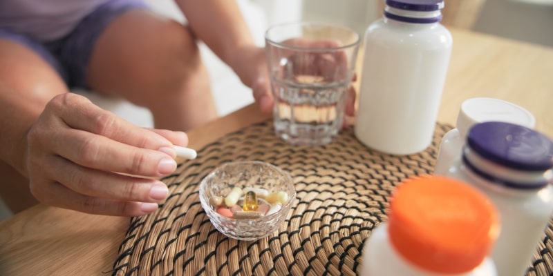 Mãos femininas pegando cápsulas em um pote de vidro sobre uma mesa com caixas de suplementos para longevidade.