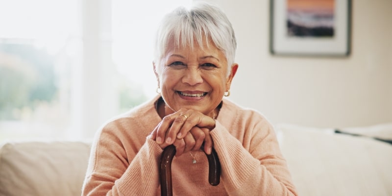 Uma mulher idosa, sentada e segurando uma bengala, sorrindo e olhando diretamente para nós. Imagem para ilustrar a matéria sobre bem-estar durante o envelhecimento.