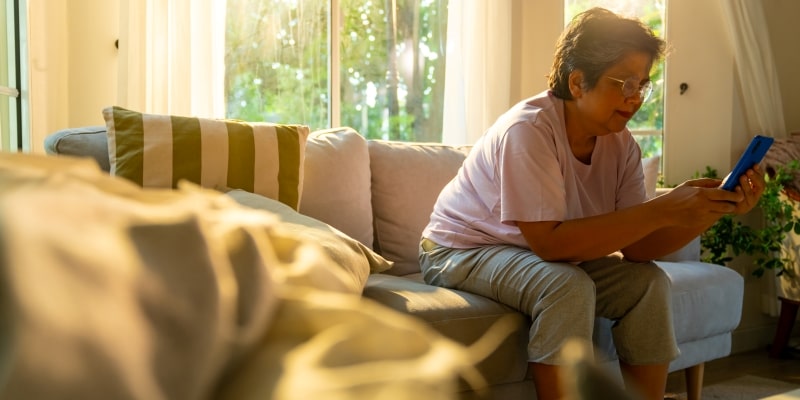 Uma senhora sentada no sofá em uma tarde com o sol de pondo e mexendo no celular para ilustrar a matéria sobre os benefícios cognitivos da tecnologia para idosos.