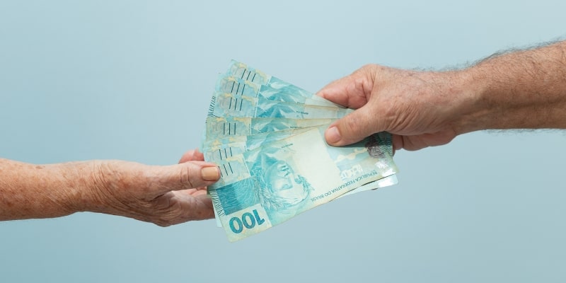Um mão entregando notas de 100 reais para outra mão, ilustrando a matéria sobre o décimo terceiro salário dos aposentados.