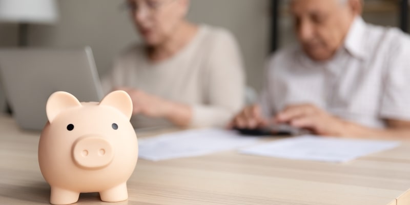 Em destaque, um cofre de porquinho, ao fundo, um casal de idosos fazendo constas e mexendo no notebook, após descobrir os direitos dos idosos garantidos por lei.