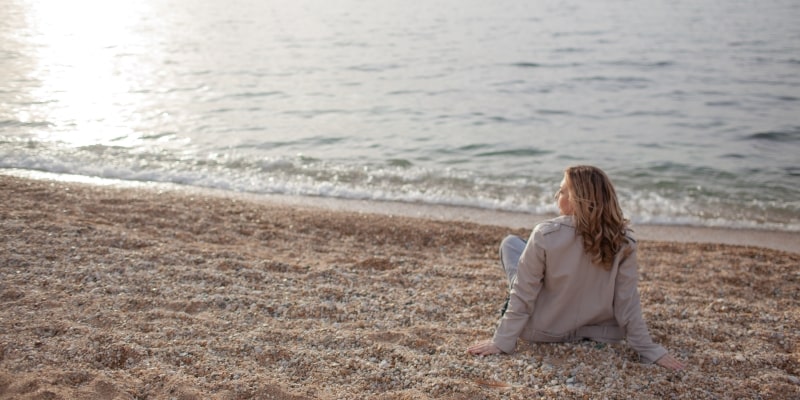 Uma mulher sentada na areia, olhando para o mar. Imagem para ilustrar a matéria sobre estar presente.