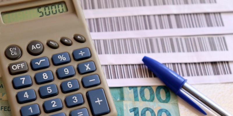 Códigos de barra de boleto, uma caneta, notas de 100 reais e uma calculadora marcando 5 mil, ilustrando a matéria sobre os principais gastos do fim de ano.