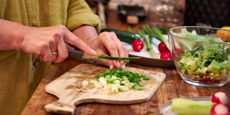 Mãos de uma mulher cortando verduras sobre uma tábua, em cima de uma mesa. Na mesa, legumes e verduras para fazer salada. Imagem para ilustrar a matéria sobre os alimentos que aumentam a longevidade.