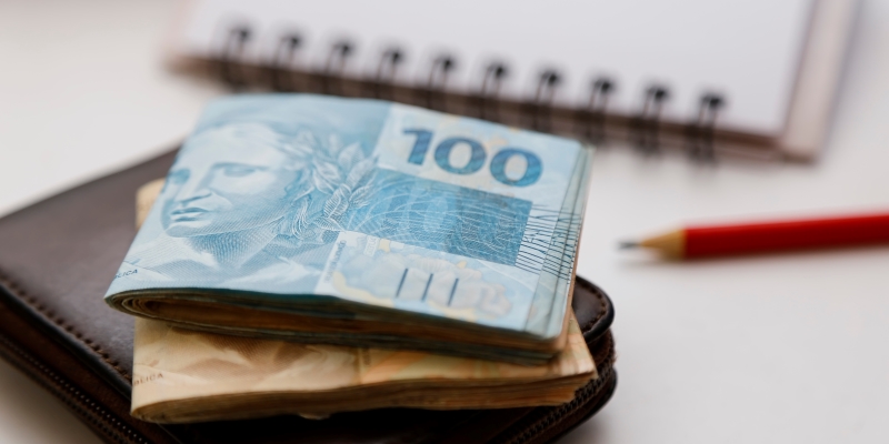 Sobre uma mesa estão um lápis, um caderno, uma carteira e dinheiro dobrado ao meio. Imagem para ilustrar a matéria sobre o conhecimento financeiro dos brasileiros.