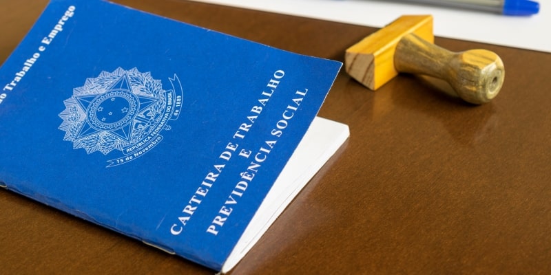 Sobre uma mesa estão uma carteira de trabalho, um carimbo e uma caneta. Imagem para ilustrar a matéria sobre o fim da escala 6x1.