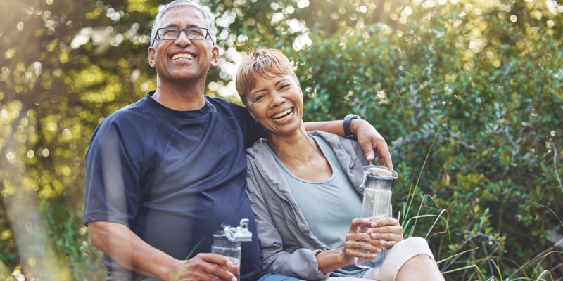 Um casal com roupa de atividade física, sentados em um lugar com arbustos, cada um com uma garrafa de água, sorrindo. Imagem para ilustrar a matéria sobre hábitos diários.