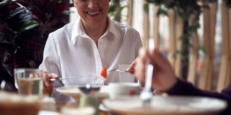  em uma senhora idosa e feliz comendo salada em um café enquanto se reúne com um amigo. Imagem para ilustrar a matéria sobre mastigação.