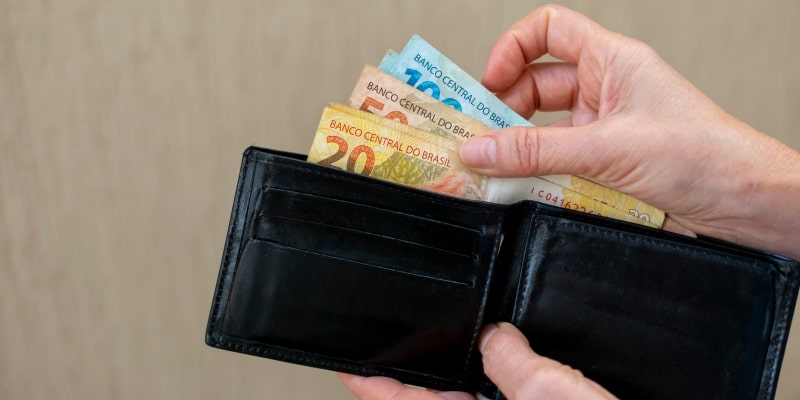 Mãos de uma pessoa tirando dinheiro de uma carteira de couro. Imagem para ilustrar a matéria sobre o novo salário mínimo 2026.
