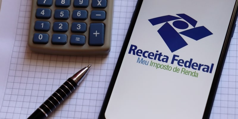 Celular com uma logo da Receita Federal ao lado de uma calculadora e de uma caneta, sobre um caderno, ilustrando a matéria sobre desconto do Imposto de Renda para aposentados do INSS.