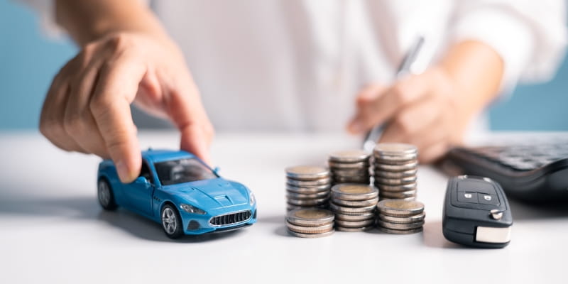 Mãos masculinas segurando uma miniatura de carro e uma caneta. Sobre uma mesa estão uma calculadora, pilha de moedas e chave de carro. Imagem para ilustrar a matéria sobre o IPVA 2026 no Nordeste.