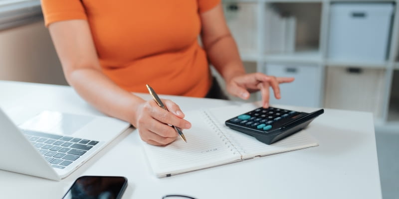 Mãos de uma mulher que está sentada em um escritório fazendo anotações em um caderno enquanto faz cálculos em uma calculadora sobre o nova isenção do Imposto do Renda.