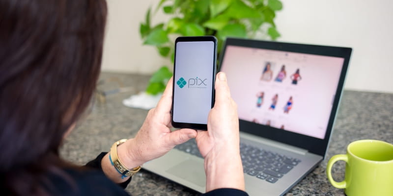 Um mulher segurando um celular com o ícone do Pix. Ao fundo, um computador mostrando produtos. Imagem para ilustrar a matéria sobre o Pix Parcelado.