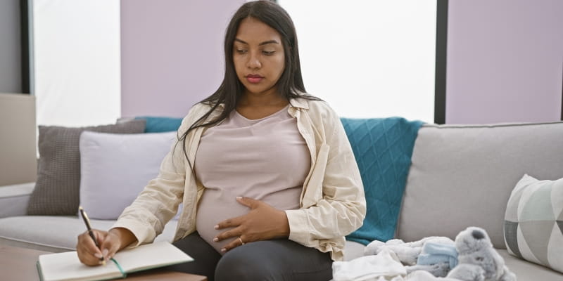 Um mulher grávida, sentada em um sofá, fazendo anotações em um caderno para o seu planejamento financeiro durante a gestação. Ao seu lado, no sofá, está um ursinho de pelúcia.