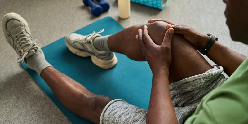 Homem negro sênior sentado no tapete de exercício segurando o joelho com as duas mãos, alongando os músculos das pernas após o treino. Imagem para ilustrar a matéria sobre como saber se estou envelhecendo bem.