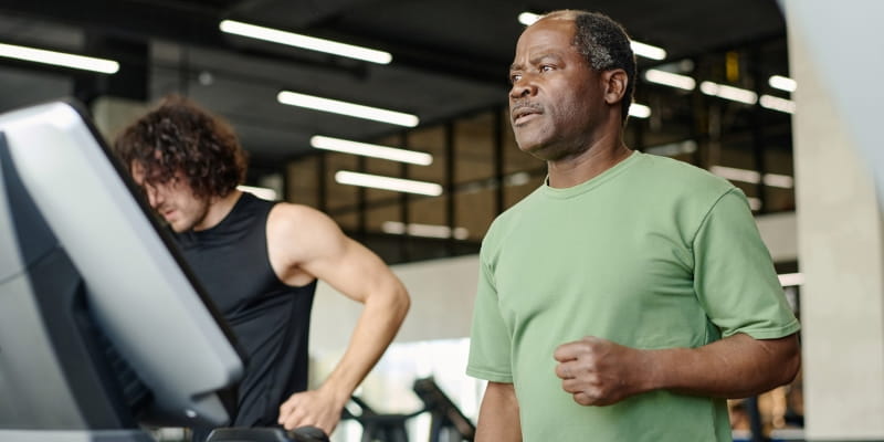 Homens, sendo um sênior negro e um jovem branco, em uma esteira de academia. Imagem para ilustrar a matéria sobre exercícios para longevidade.