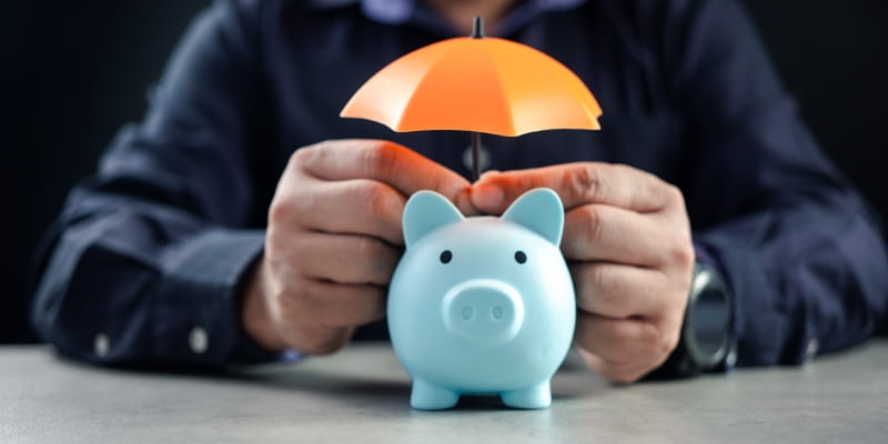 Mãos masculinas cobrindo um cofrinho com um guarda-chuva. Imagem para ilustrar a matéria sobre previdência privada 2026.