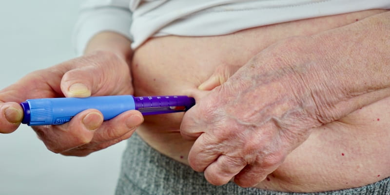 Foco das mãos de uma pessoa idosa aplicando o remédio na barriga ao ilustrar o uso de canetas emagrecedoras por idosos.