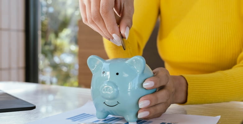 Mãos de uma mulher colocando dinheiro em um cofre de porquinho, ilustrando a matéria sobre como economizar dinheiro em 2026.