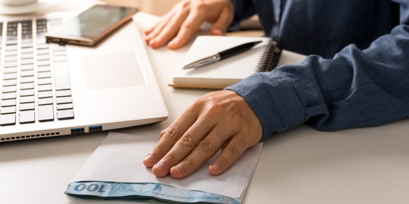 Mãos de um homem segurando um envelope de dinheiro. Ele está em uma mesa de computador com uma caneta, um laptop, um caderno e uma caneta. Imagem para ilustrar a matéria sobre como planejar aposentadoria de 10 mil reais.