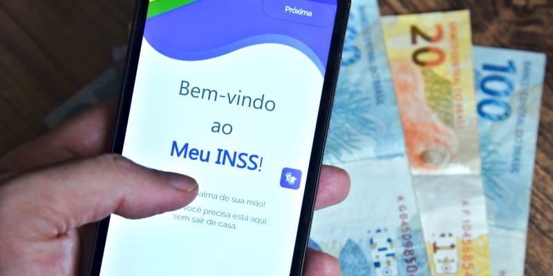 Uma mão segurando o celular que está com o aplicativo do Meu INSS aberto e, ao fundo, notas de dinheiro, ilustrando a matéria sobre como simular aposentadoria pelo Meu INSS.