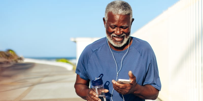 Um homem negro e idoso, ouvindo música com fone de ouvido, enquanto caminha em área aberta. Ele olha para o celular e carrega uma garrafa d'água. Imagem para ilustrar a matéria sobre como ter mais longevidade.