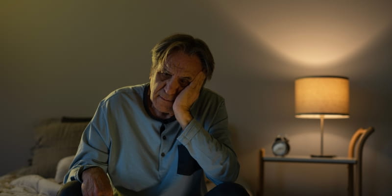 Um homem idoso, em seu quarto, com o semblante abatido após ter uma má noite de sono, sentindo as consequências de dormir pouco.