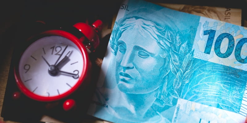 Uma nota de 100 reais ao lado de um mini relógio, ilustrando a matéria sobre a contestação dos descontos indevidos do INSS.