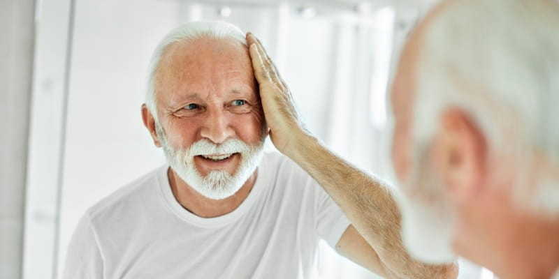 Um humem idoso com cabelos e barba branca, passando pela sua fase de envelhecimento biológico. Ele está sorrindo e passando a mão na testa enquanto se olha no espelho.