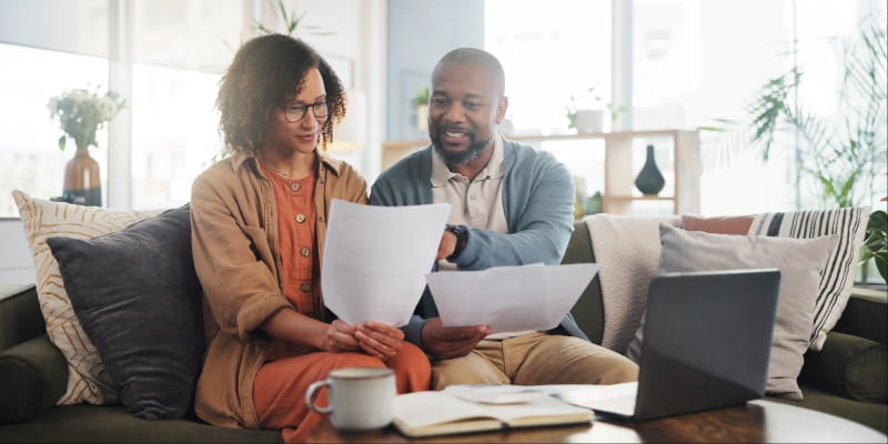 Um casal negro, com cerca de 40 anos, sentados no sofá de sua casa. A mulher segura papéis enquanto o homem aponta para ele. Na frente, há uma mesa centro com um laptop. A imagem ilustra a prática de hábitos financeiros para envelhecer bem.