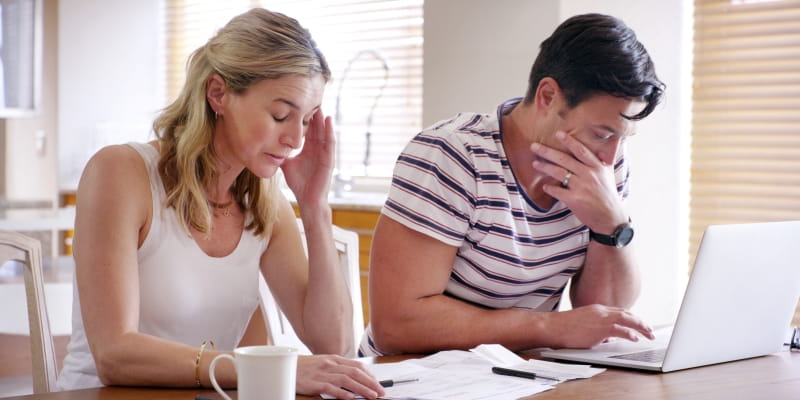 Casal estressado  em casa com um laptop e com contas e papelada, demonstrando problemas financeiros por conta de infidelidade financeira.
