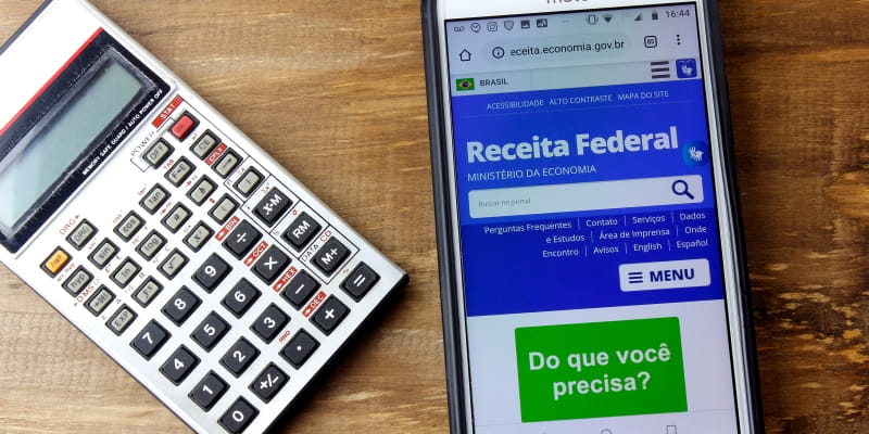 Sobre uma mesa estão uma calculadora e um celular com o site da Receita Federal aberto, ilustrando a matéria sobre o IR 2026.